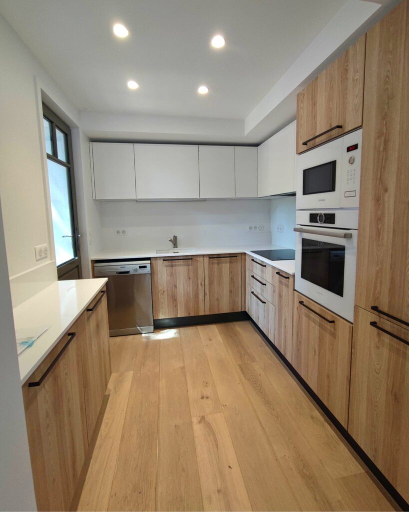 Cuisine moderne en L avec meubles bas en bois clair, placards hauts blancs, plan de travail blanc et électroménager Bosch/Siemens intégré. Sol parquet.