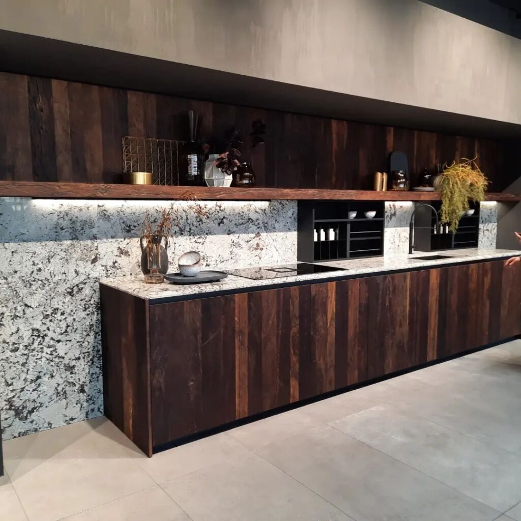 Cuisine contemporaine avec meubles bois foncé à lattes, comptoir et crédence en granit moucheté. Étagère flottante éclairée, évier noir, cuisinière et décorations.