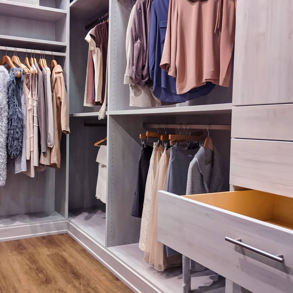 Placard dressing moderne en bois clair avec étagères et penderies. Vêtements pastel accrochés, un tiroir ouvert révèle l'intérieur en bois.