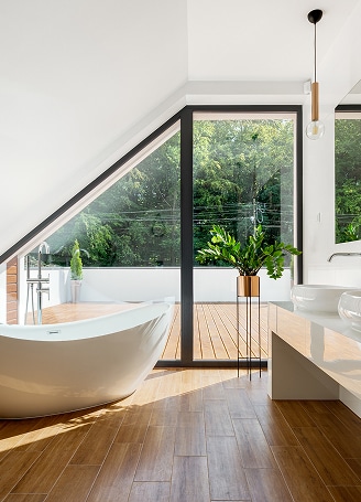 Salle de bain moderne lumineuse avec baignoire blanche sur pied, parquet en bois et grande fenêtre offrant une vue sur la nature.
