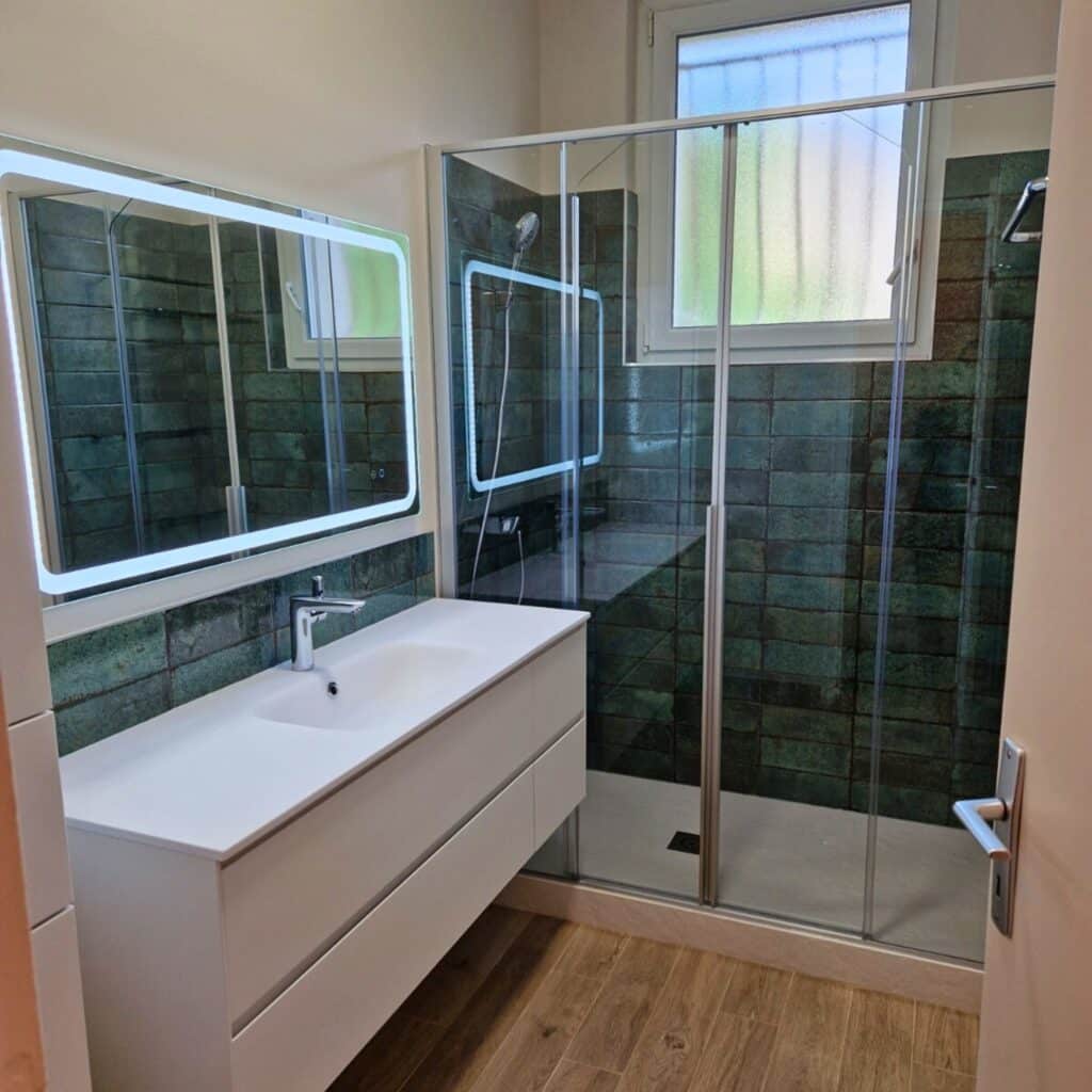 Salle de bain moderne avec meuble vasque blanc, miroir lumineux, douche à l'italienne carrelée de vert, et sol effet bois clair.